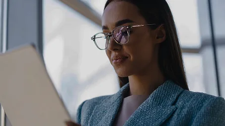 Una mujer, vestida formalmente, sale de una oficina con puertas de cristal, sosteniendo su tableta electrónica para revisar las últimas actualizaciones de su curso de Power Bi.