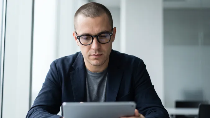Un hombre de pie hace anotaciones en un cristal mientras ve de reojo su laptop ubicada en un escritorio a su lado, donde hace seguimiento a su diplomado en línea.